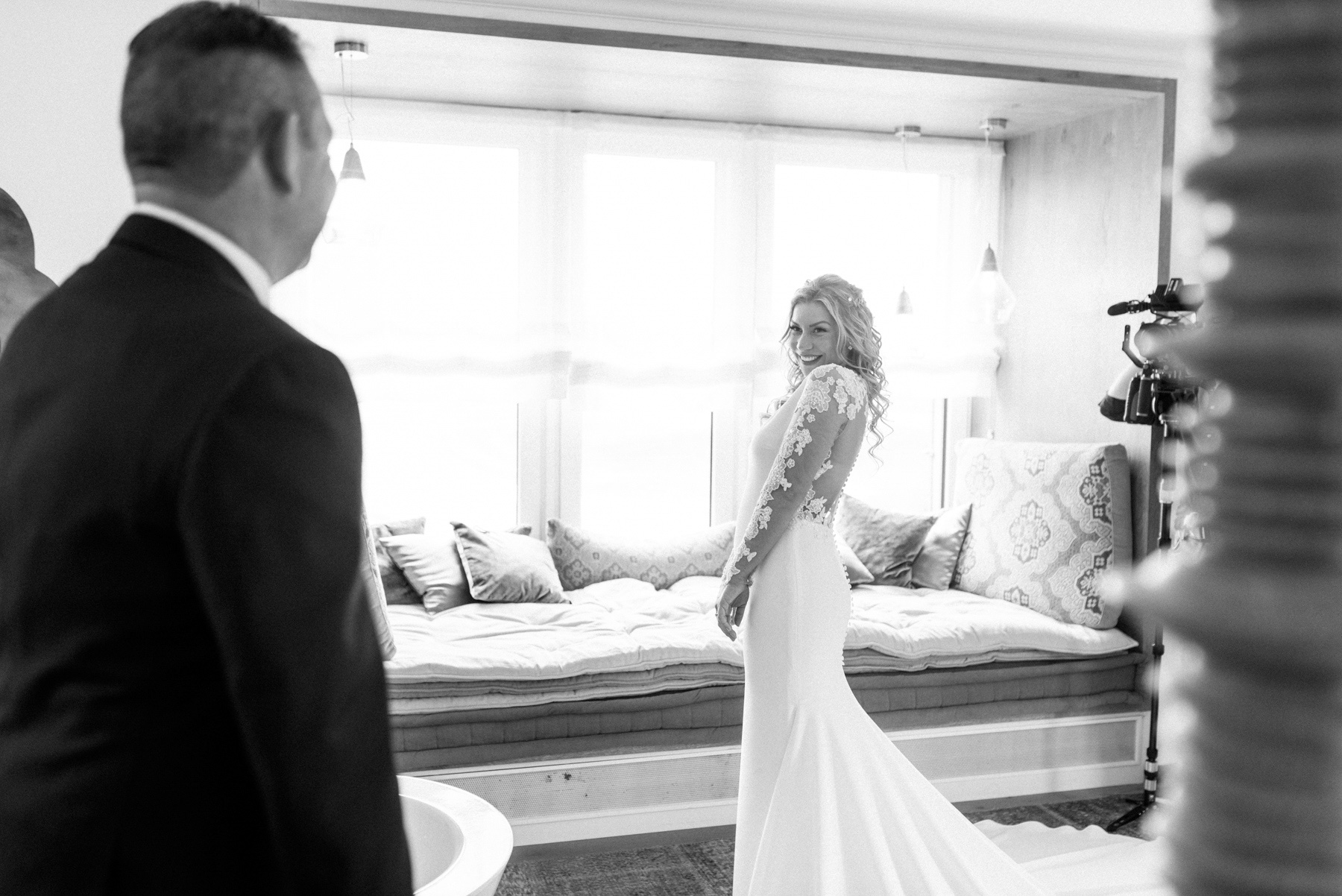 Braut im eleganten Hochzeitskleid lächelt ihrem Vater entgegen beim First Look vor einer Fensterbank mit Kissen.