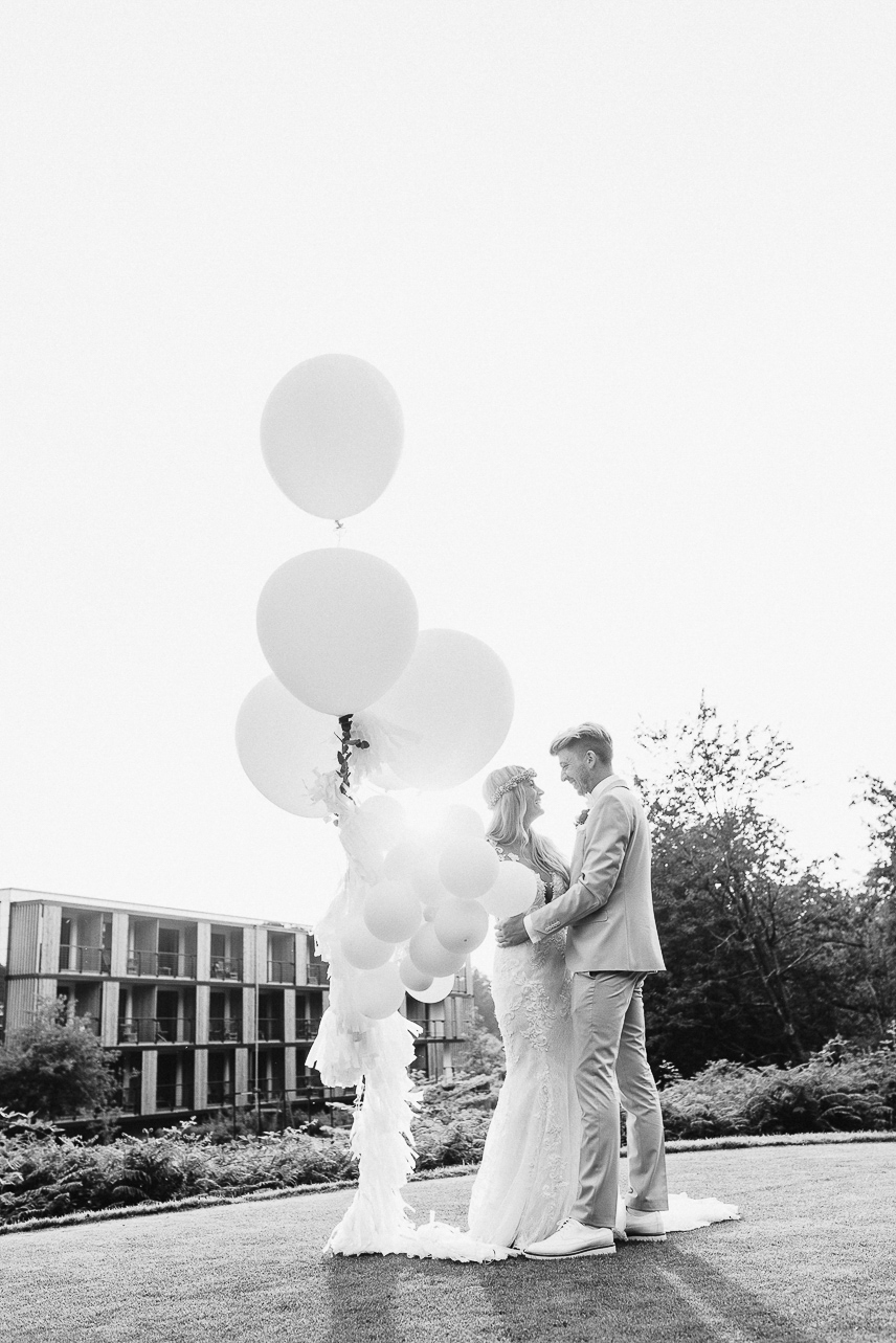 Schwarz-weißes Bild eines Brautpaares, das sich küsst, umgeben von schwebenden Luftballons auf dem Rasen – romantische und festliche Hochzeitsfotografie.
