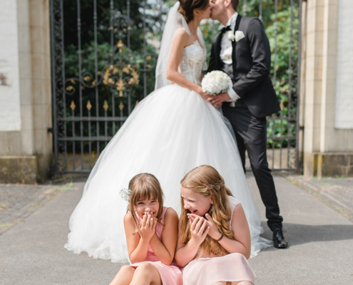Romantische Hochzeitsfotografie von Jeean Hochzeitsfotografie – Das Brautpaar küsst sich vor einem großen Tor, während die Blumenkinder beschämt und süß auf dem Boden sitzen und zuschauen.