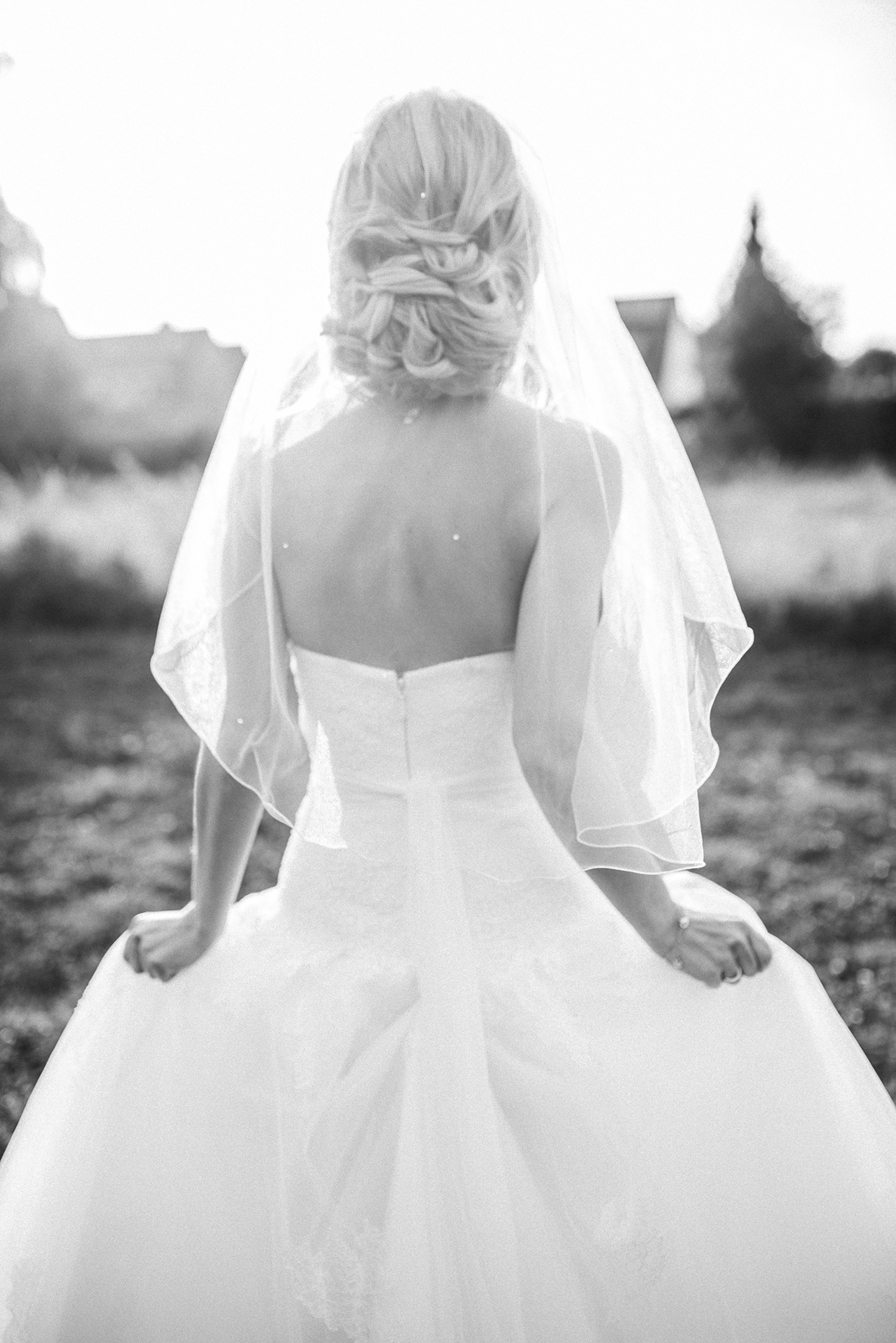 Schwarz-Weiß-Fotografie von Jeean Hochzeitsfotografie – Die Braut hält ihr Brautkleid von hinten leicht hoch, eingefangen in einem eleganten Moment im Grünen.