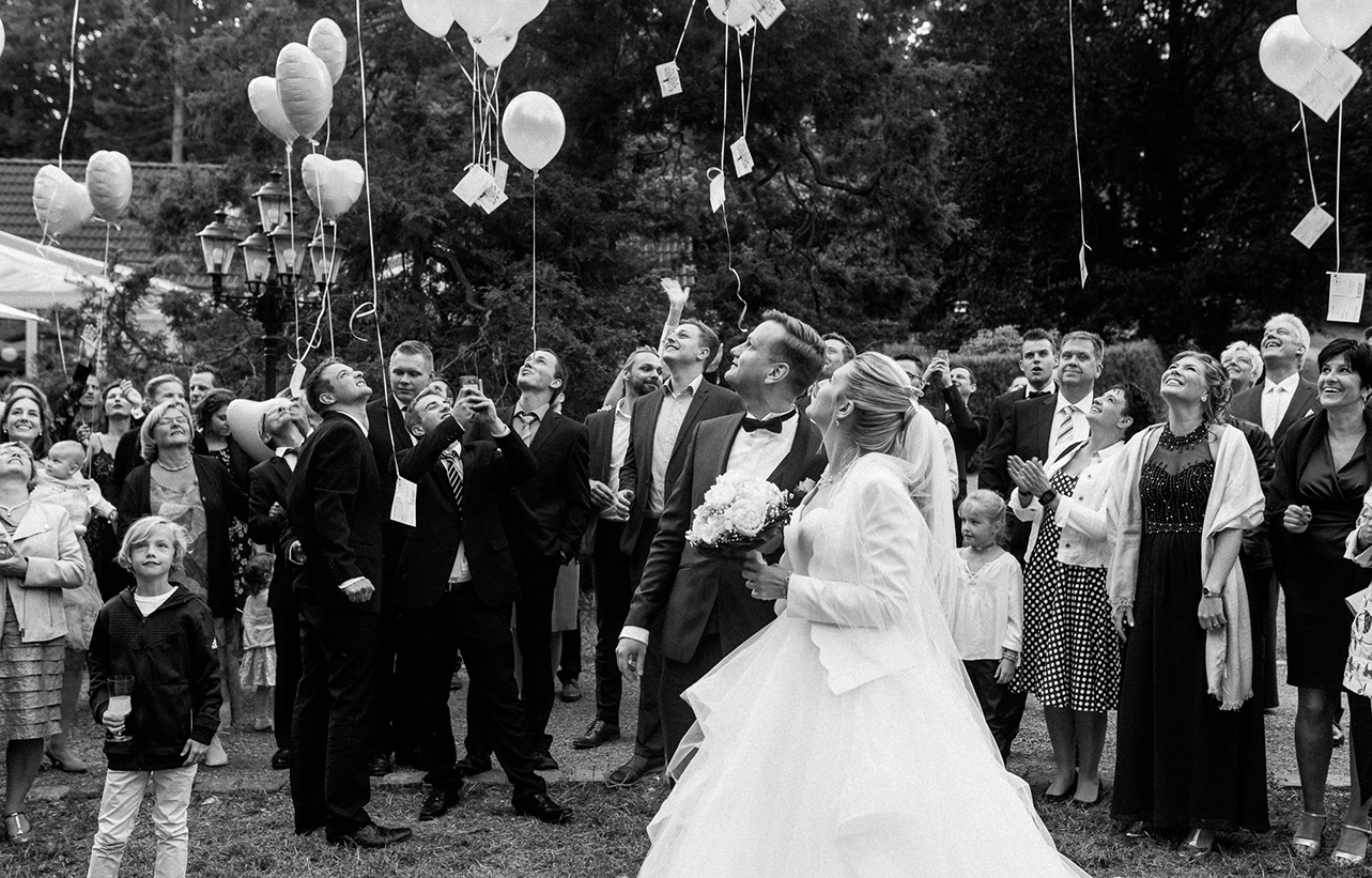 Hochzeitsfotografie von Jeean Hochzeitsfotografie – Alle Gäste lassen Luftballons mit Karten hochsteigen, während sie in den Himmel schauen, das Brautpaar wird seitlich abfotografiert.