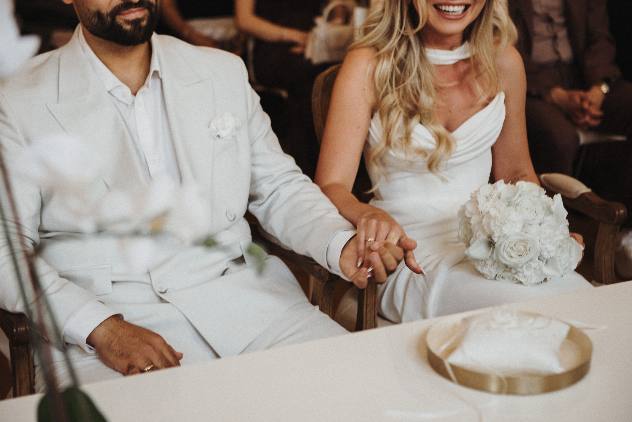 Braut und Bräutigam in weißen Outfits halten bei ihrer Hochzeit Händchen, während die Braut einen weißen Blumenstrauß auf dem Schoß trägt.