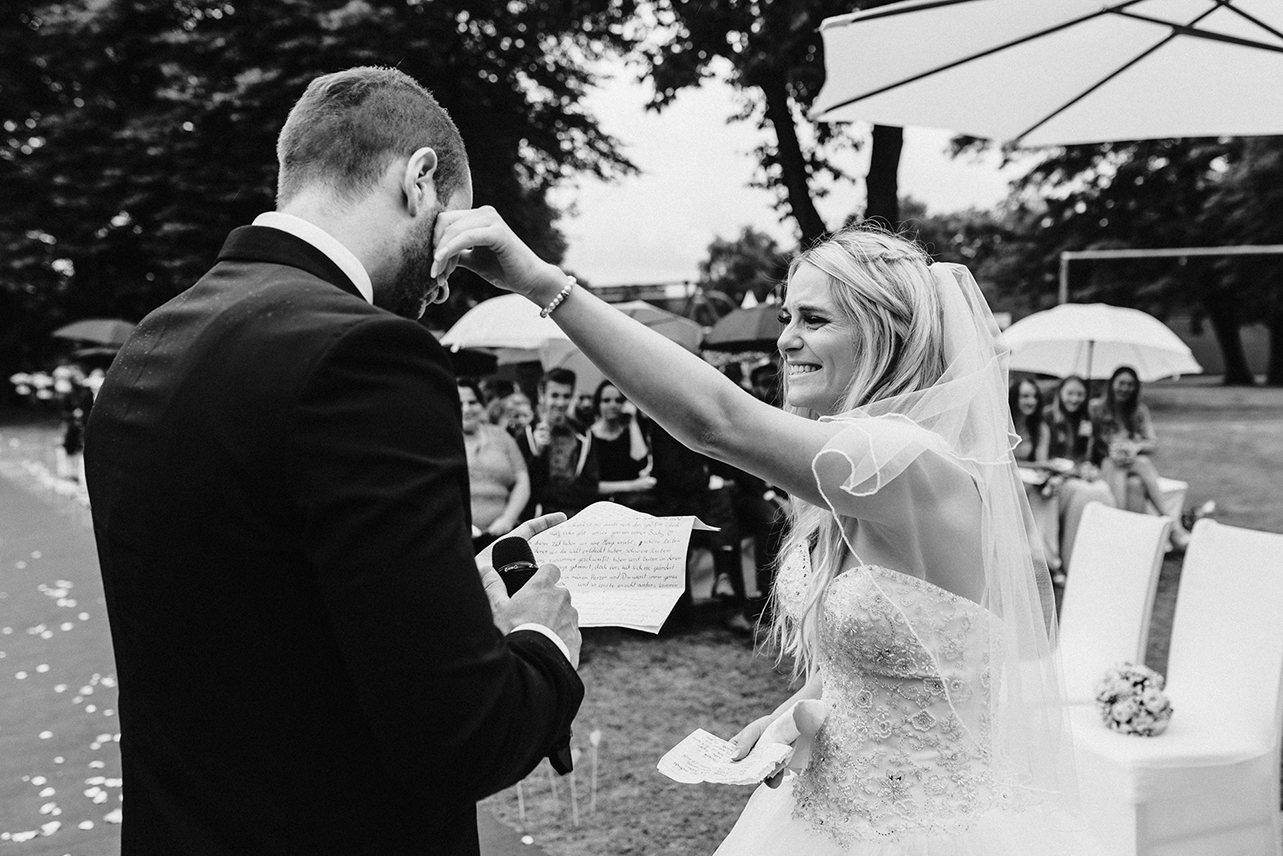 Emotionale Hochzeitsfotografie von Jeean Hochzeitsfotografie – Die Braut streicht dem Bräutigam liebevoll die Tränen vom Gesicht während der Zeremonie im Garten.