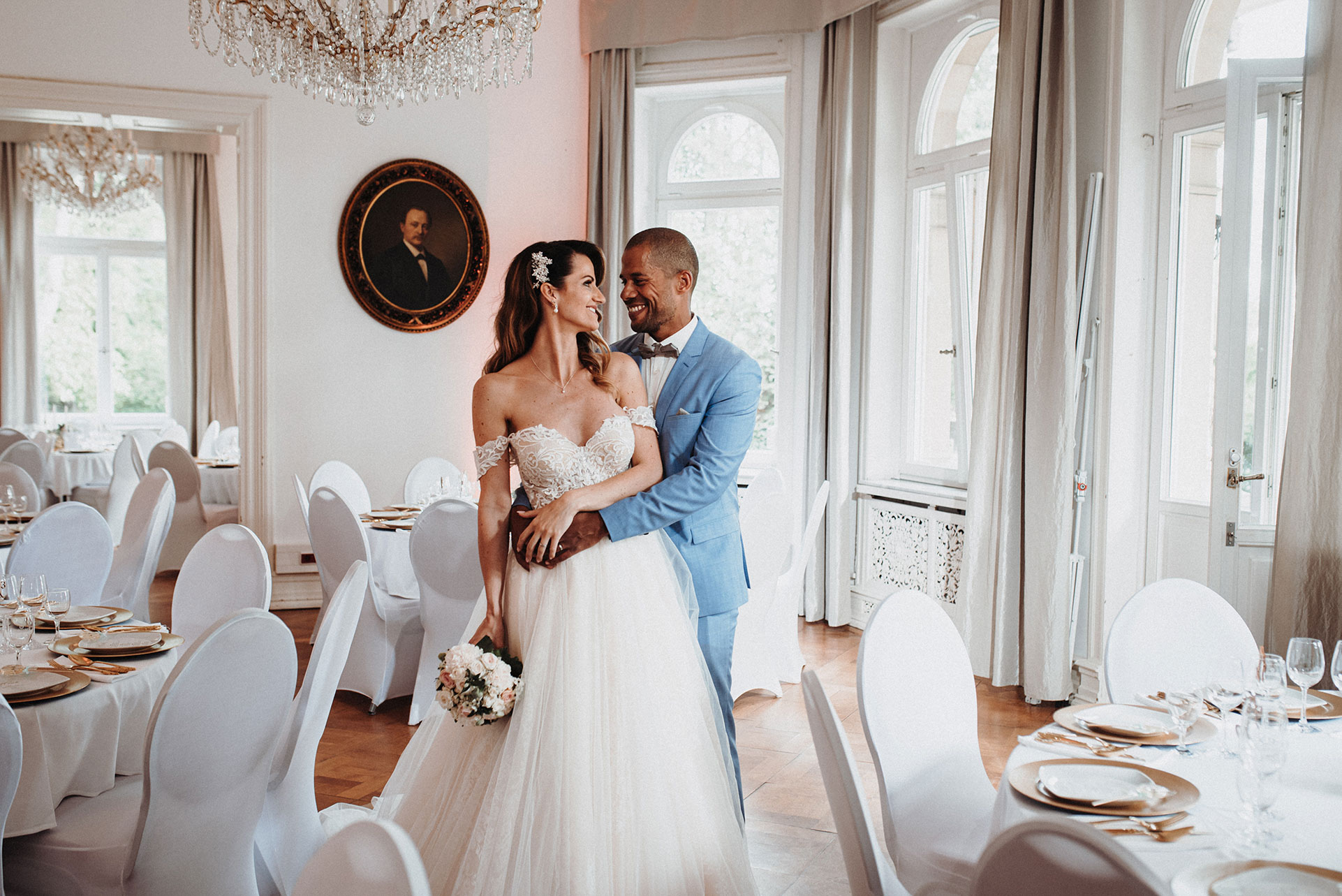 Hochzeitspaar in elegantem Saal mit Kronleuchtern, der Bräutigam umarmt die Braut liebevoll