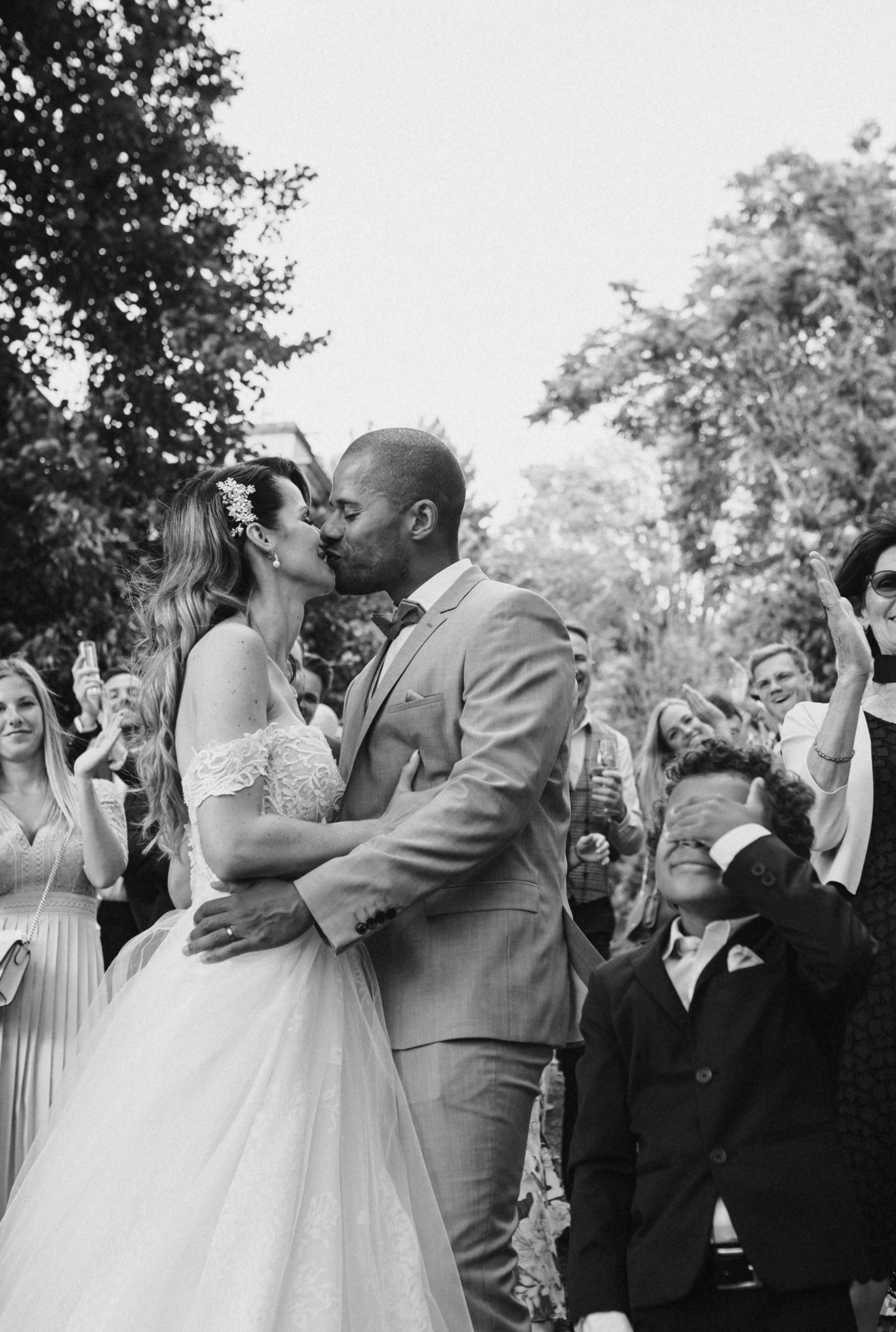Hochzeitspaar küsst sich umgeben von jubelnden Gästen, während ein kleiner Junge sich spielerisch die Augen zuhält.